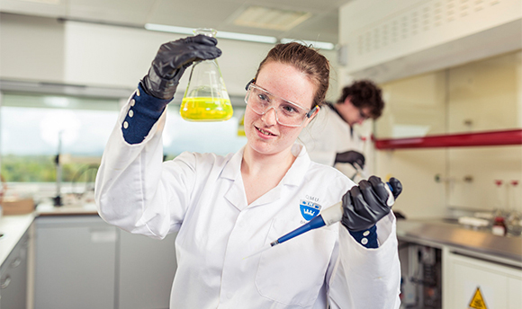A 6Ͽ student wearing PPE and holding up a beaker of yellow liquid