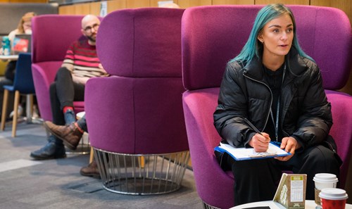 Student with vibrant blue hair sat on purple pod seat in 6Ͽ Atrium